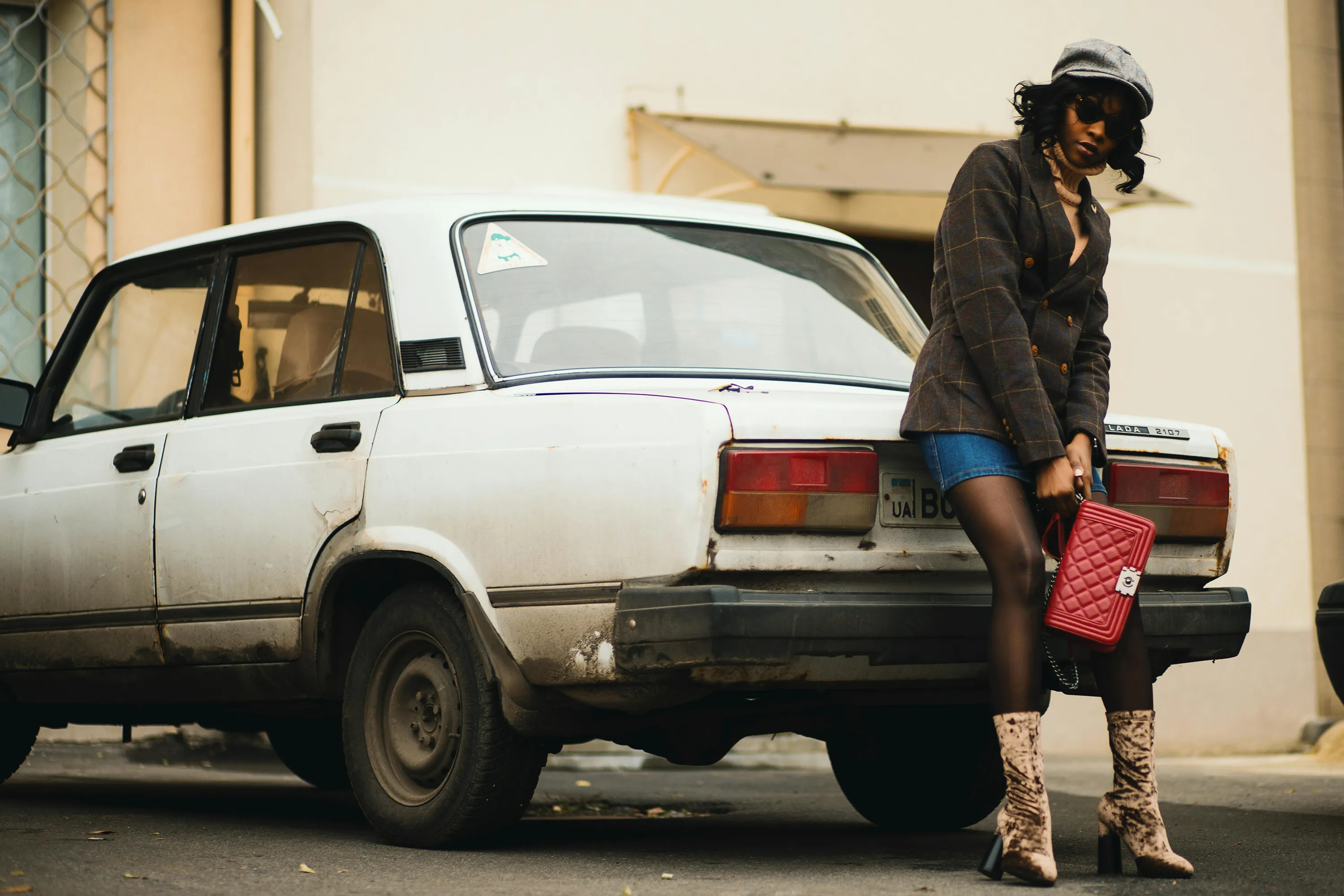A stylish woman in a blazer and snakeskin boots leans against an old white car