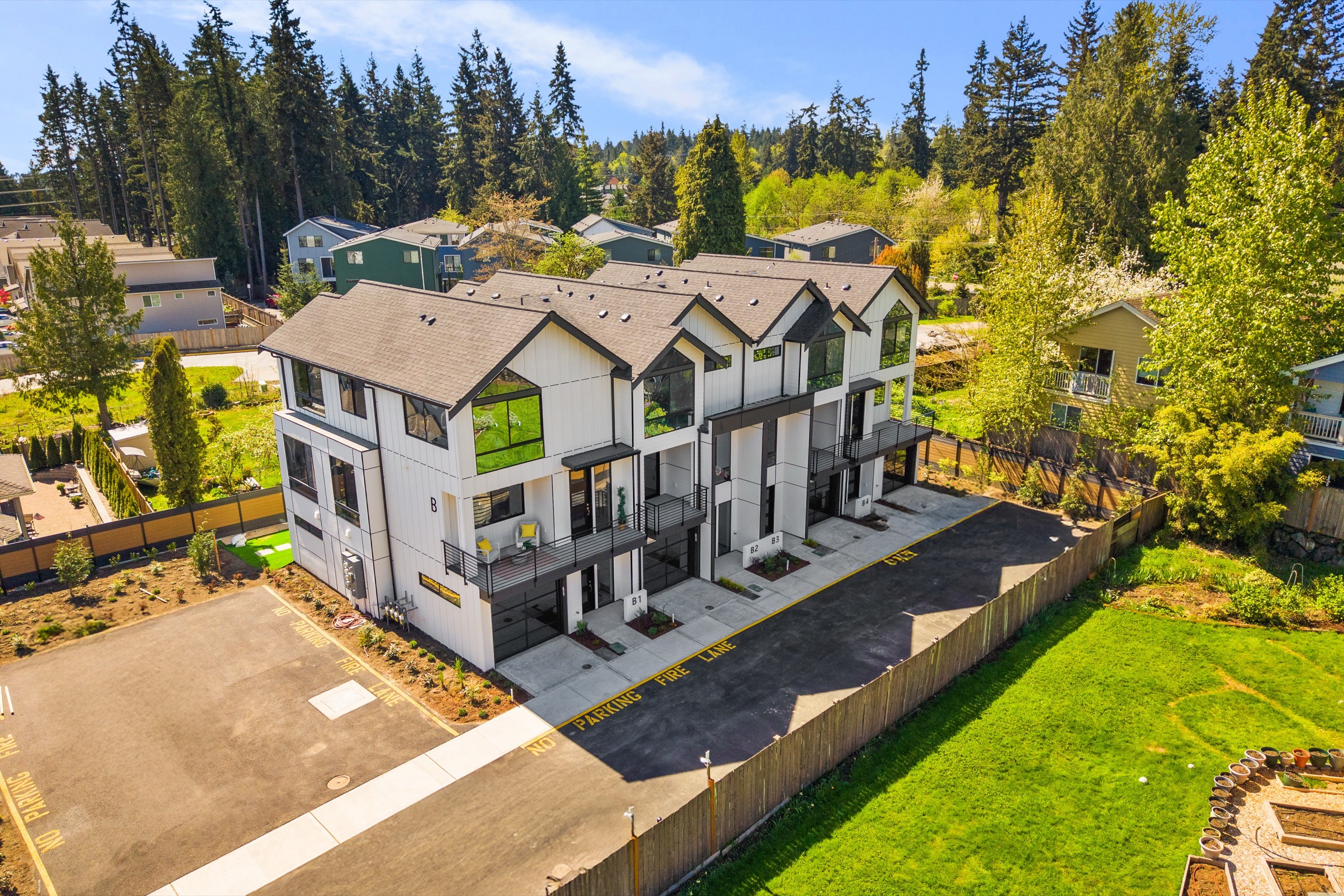 Aerial view of a newly built luxury townhome complex with grey roofing, private balconies, manicured landscaping, and mature evergreen trees in a Pacific Northwest suburb