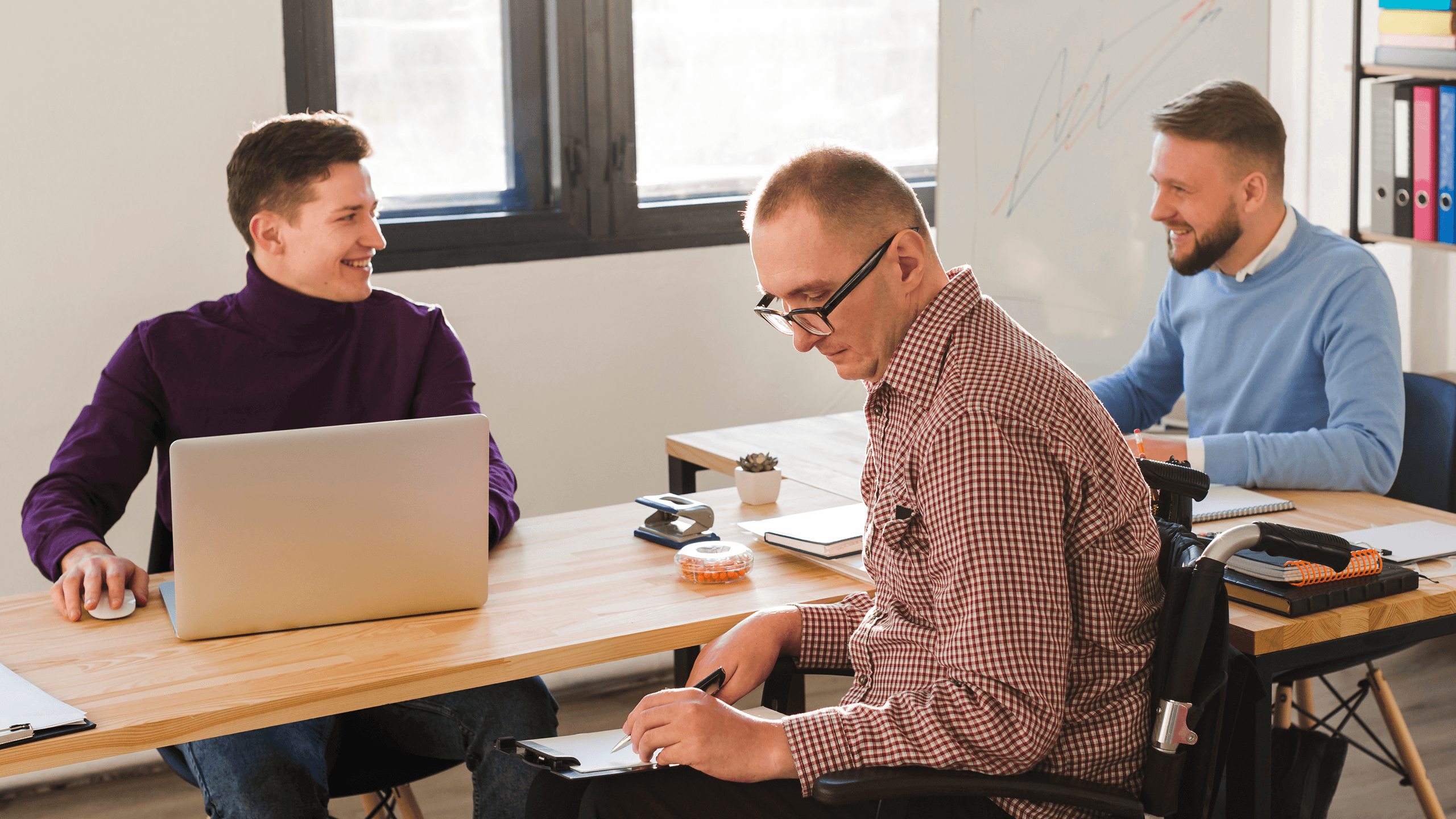 Three coworkers in a meeting, with one using a laptop and another taking notes at a desk.