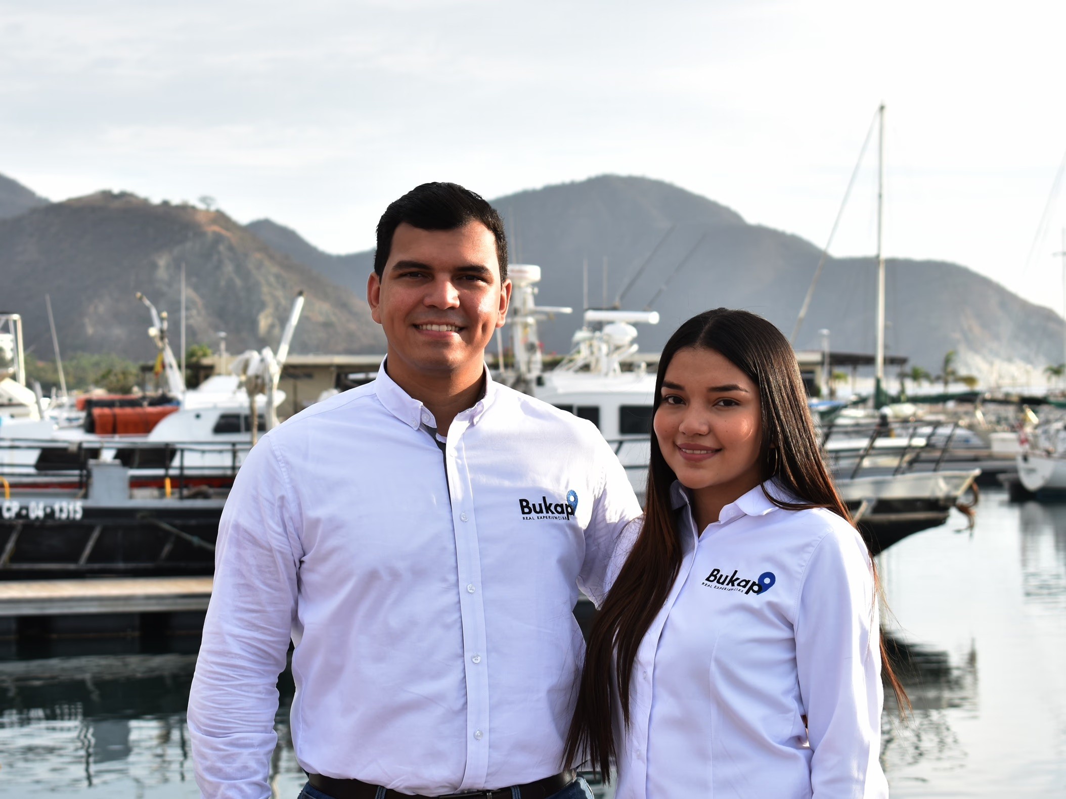 A man and woman in white "Bukap" branded shirts smiling in a harbor with boats and mountains.
