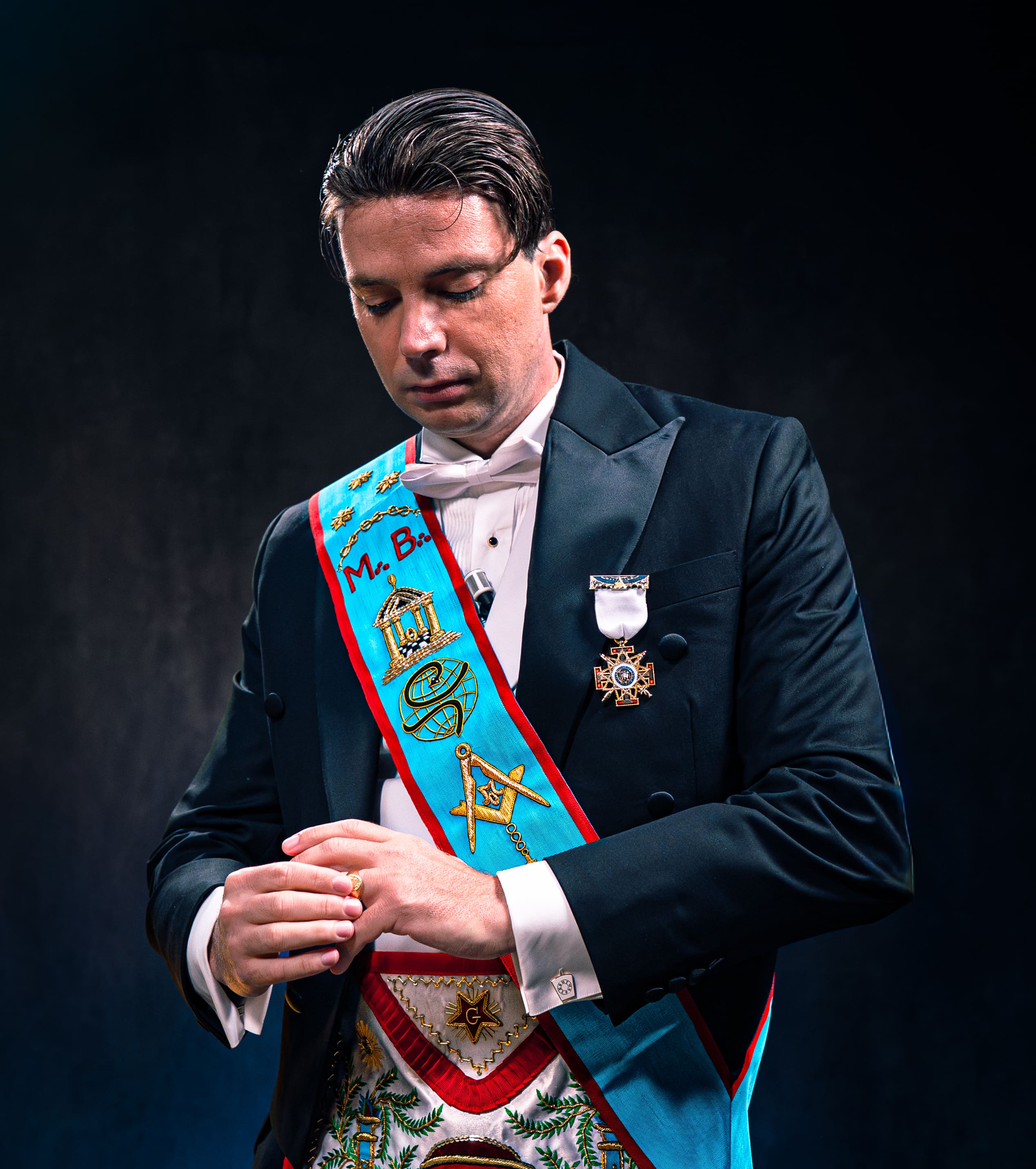 Photographie de portrait professionnelle d'un homme en tenue de Franc-Maçonnerie, portant un smoking noir, un nœud papillon blanc et des insignes. L'homme est vu de face, les yeux baissés et les mains ajustant une bague. L'accent est mis sur l'écharpe d'officier bleu vif (sautoir) ornée de symboles maçonniques (équerre et compas, temple). Le fond sombre et l'éclairage dramatique créent une atmosphère sérieuse et mystérieuse. Cette photo de haute qualité, optimisée par Nux-Studio, est un exemple de production photo pour des interviews sur l'histoire, les organisations secrètes ou des sujets de société (Franc-Maçonnerie).