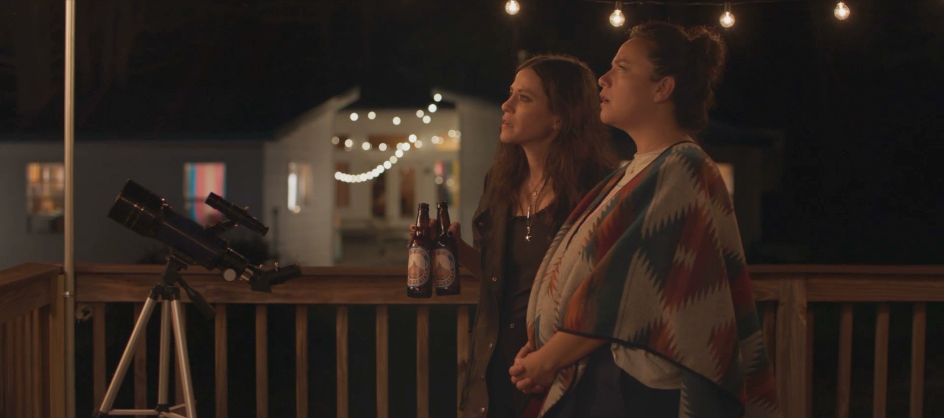 Still from The Floaters: Two women standing on a porch at night, gazing up at the sky. One holds two bottles of beer, while a telescope sits nearby.