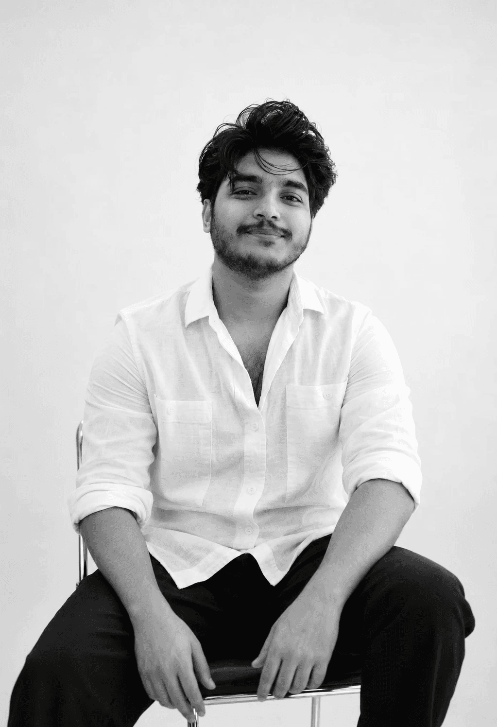 Casual studio portrait of a bearded man with curly hair in a white shirt, arms crossed, on a light gray background.