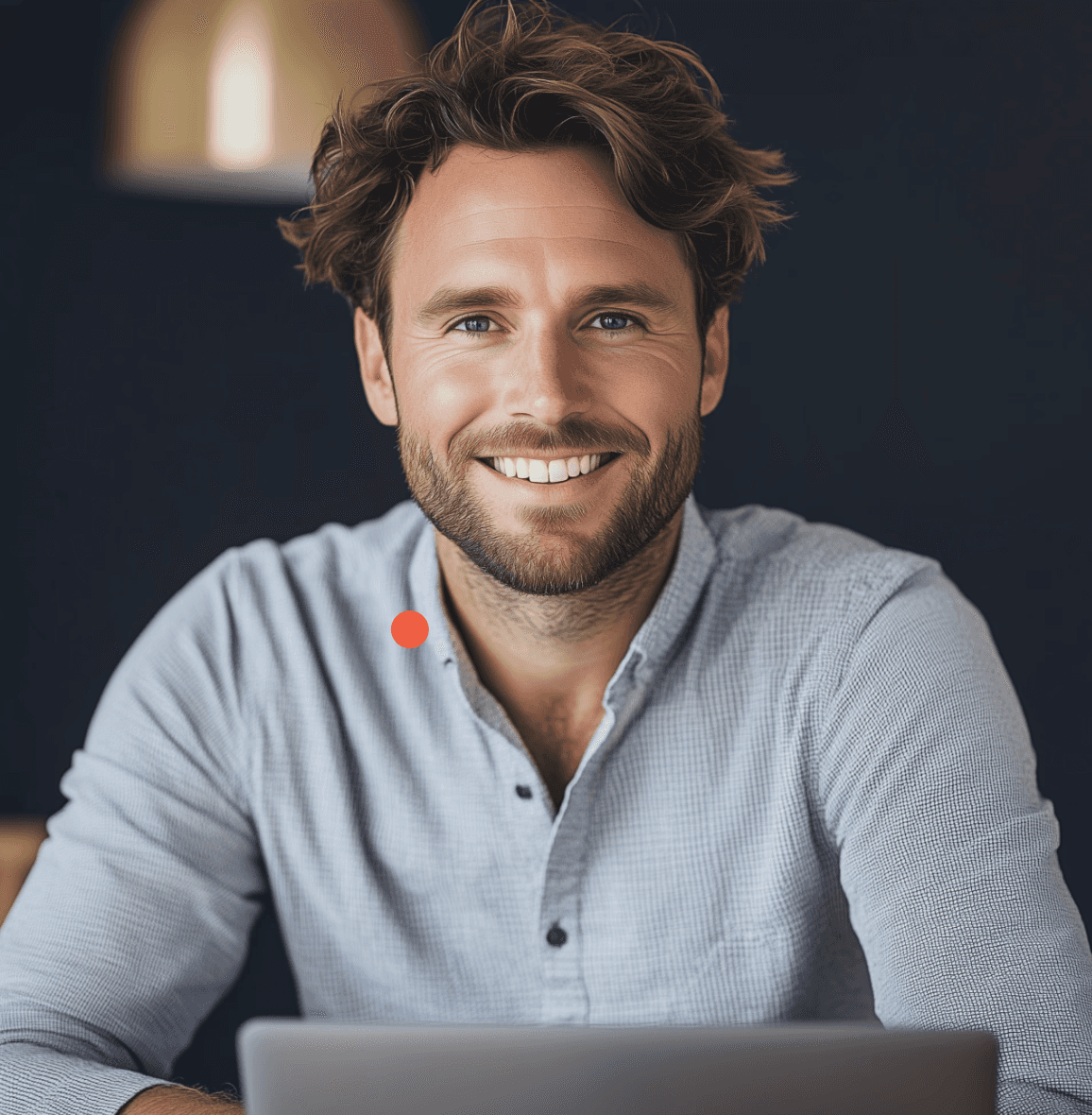 A smiling man with curly hair is sitting at a desk, looking at the camera, with a laptop and a lamp in the background.