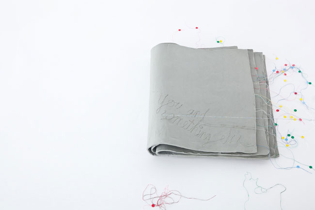 A gray fabric roll lies on a white surface. The words "You and something else" are written in black marker on the fabric. Colorful thread is scattered around the roll and across the surface.