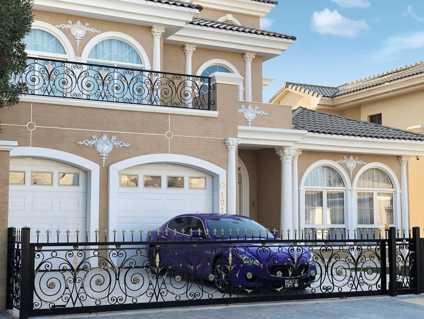 A sleek, purple luxury car is parked in front of a large, elegant two-story house featuring ornate white columns, intricate wrought-iron railings, and multiple arched windows, all under a clear blue sky.