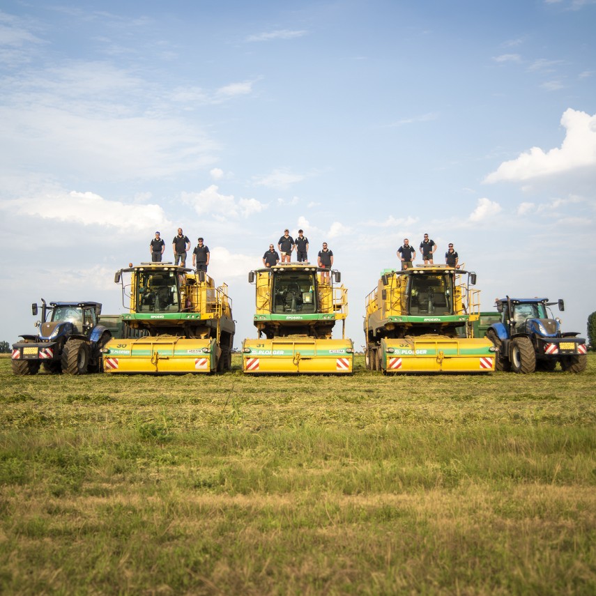 A harvest team posing on top of three numbered Ploeger pea harvesters (30, 31 and 32) lined up in a field, flanked by two blue New Holland tractors, under a sunny sky with white clouds.