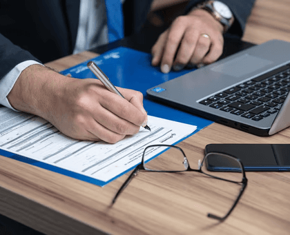 Person in a suit filling out paperwork at a desk with a laptop, phone, and glasses.
