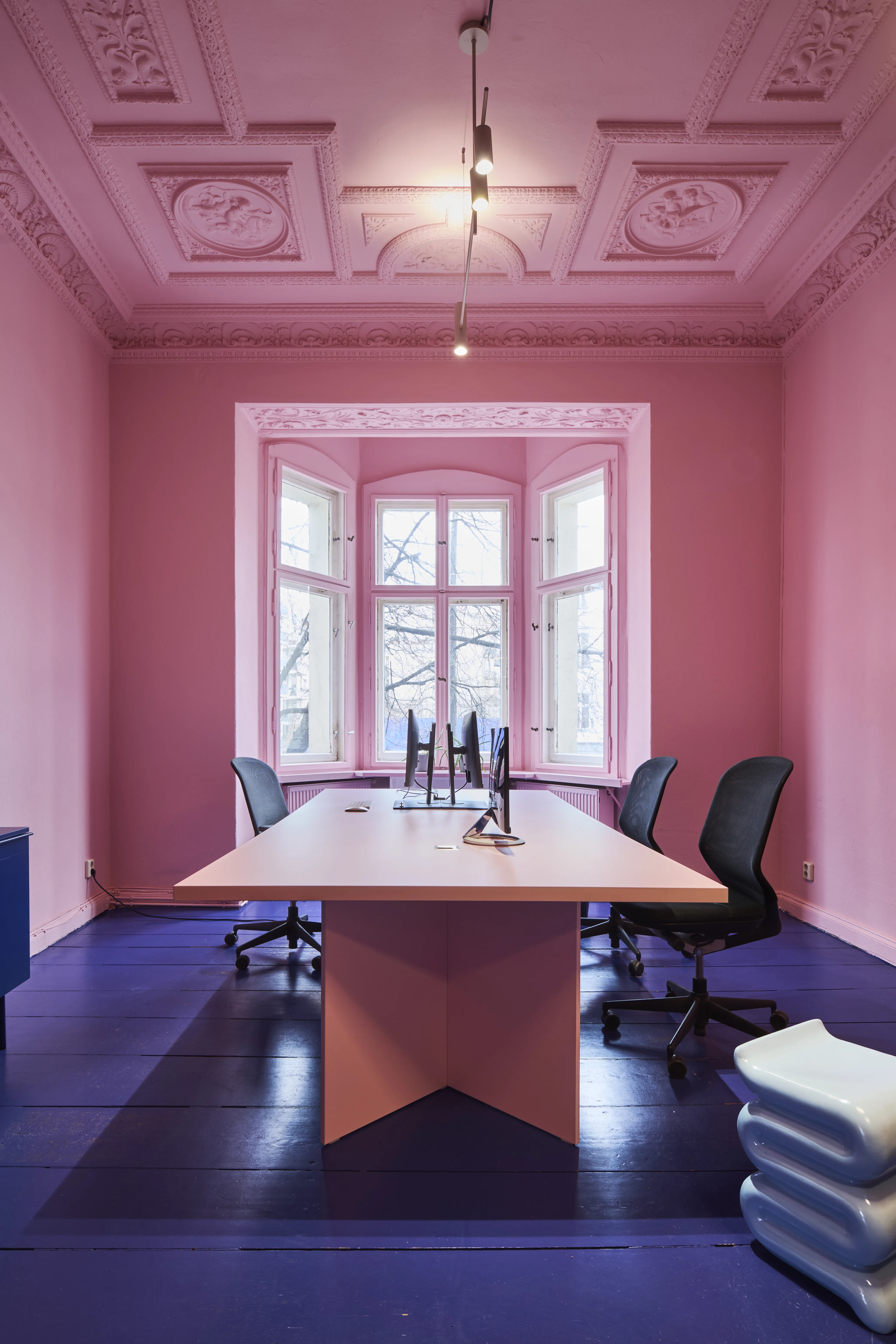 Pink office with ornate stucco ceiling, rose desk, black chairs and dark blue wooden floor.