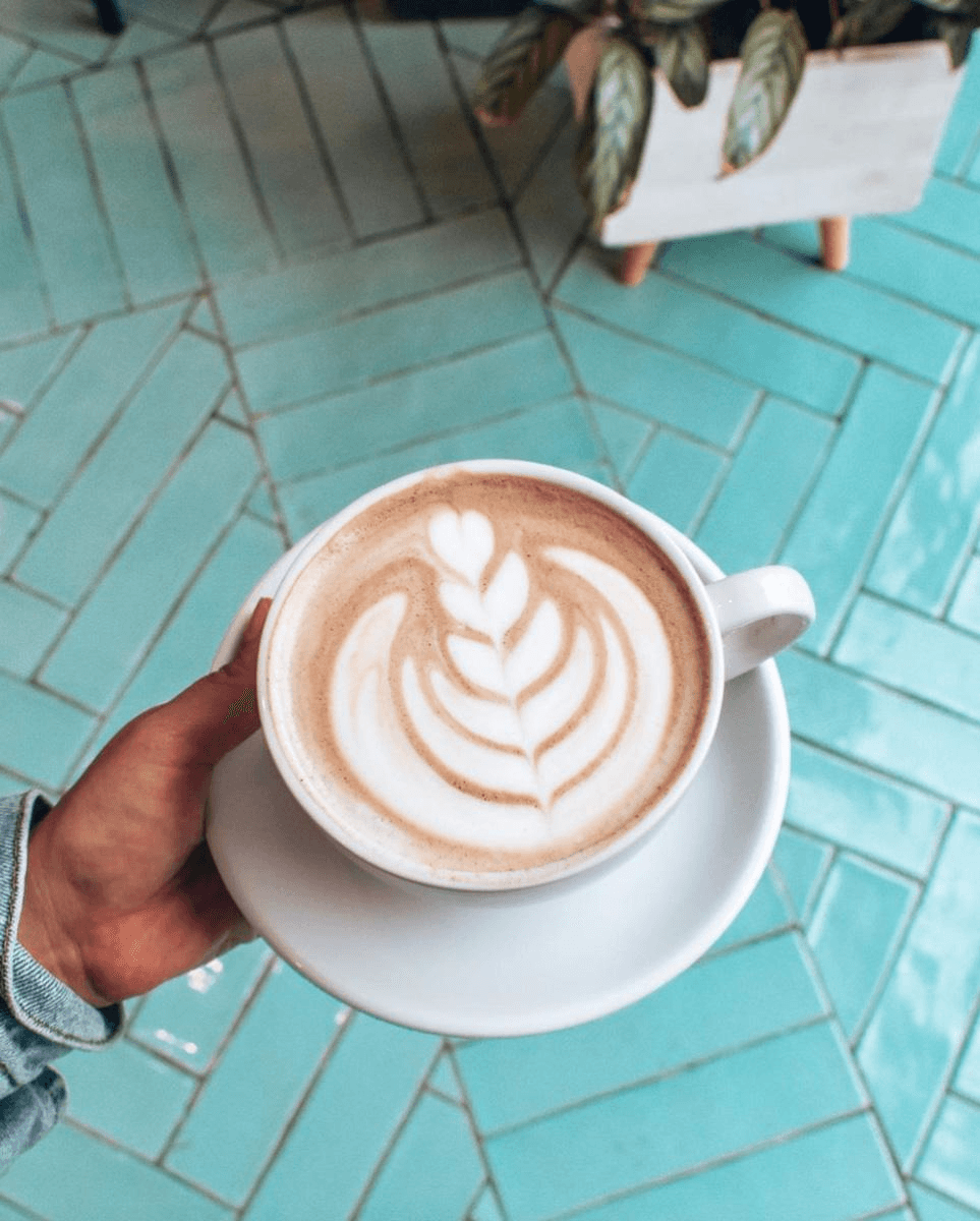 A hand holds a white cup of creamy latte art coffee on a white saucer, set against a teal herringbone-tiled floor, with a glimpse of a potted plant in the corner.