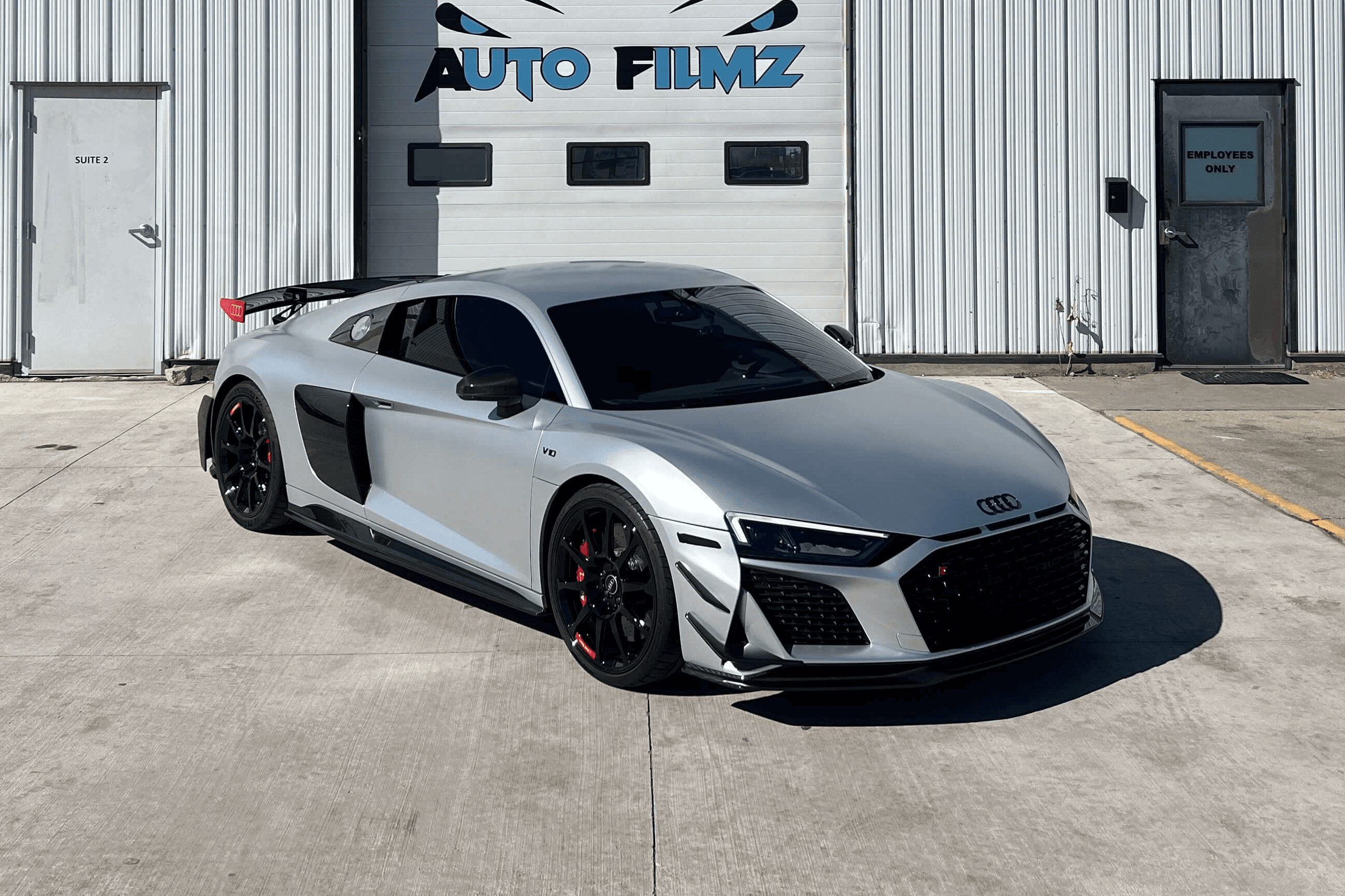 A sleek silver Audi R8 sports car is parked in front of a white industrial building with the sign "Custom Auto Filmz" displayed prominently above one of the garage doors.