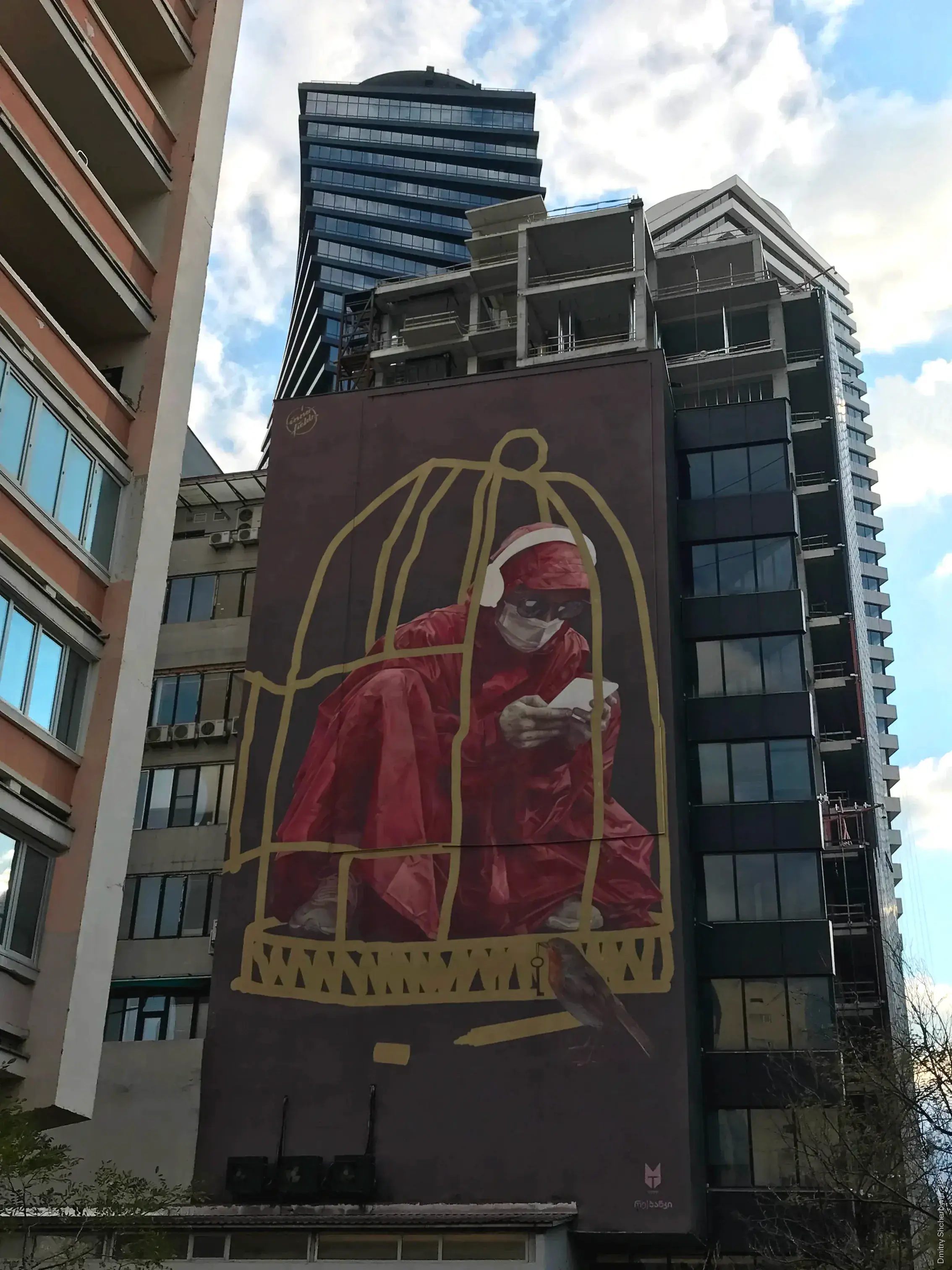 A tall building mural depicting a person wearing a red hooded garment, a white face mask and sitting inside a hand-drawn bird cage, looking down at a phone in their hands. At the bottom of the cage sits a small bird with a key in its beak.