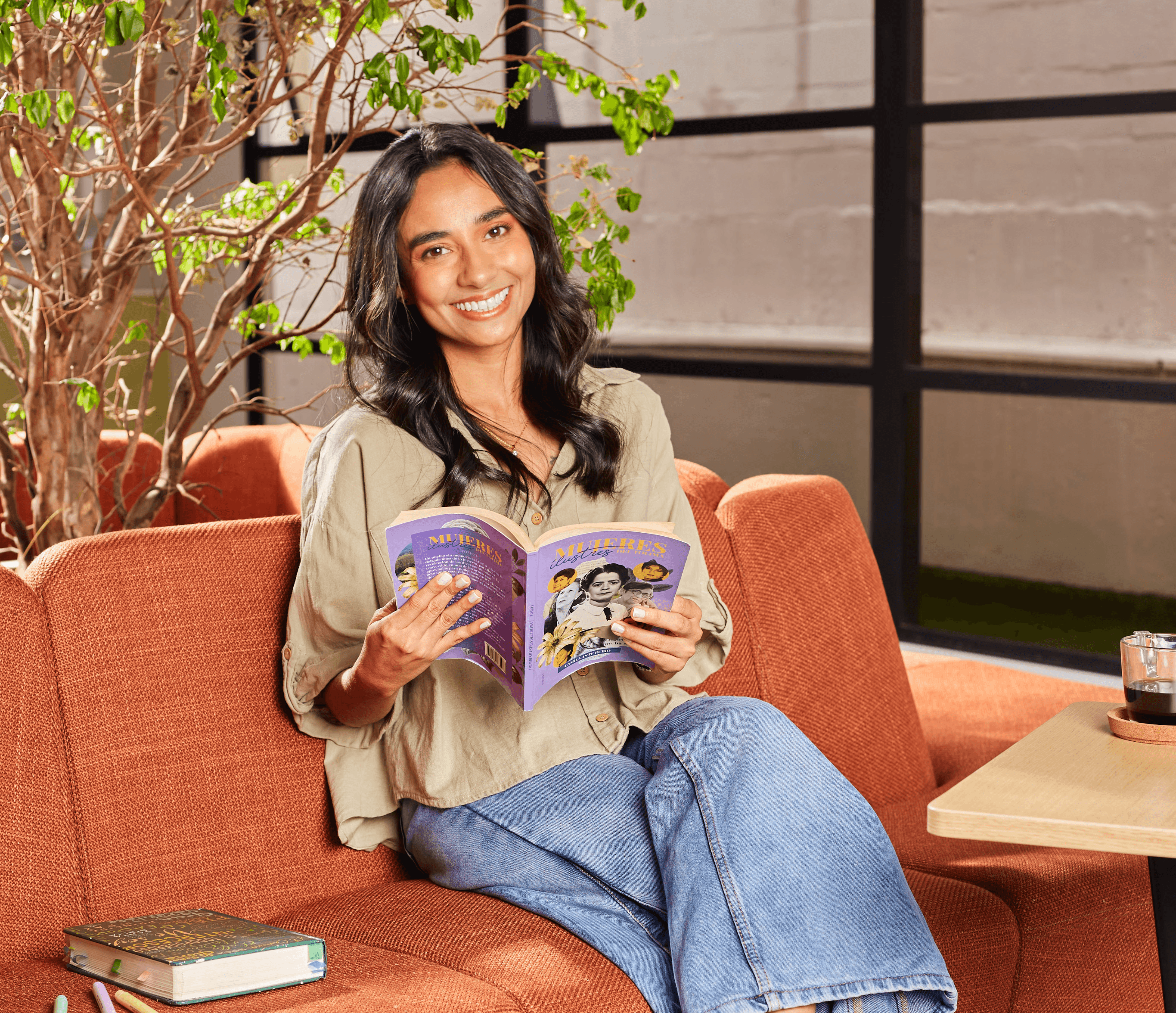 Retrato de la autora sonriendo, sentada cómodamente en un sofá naranja y leyendo un libro