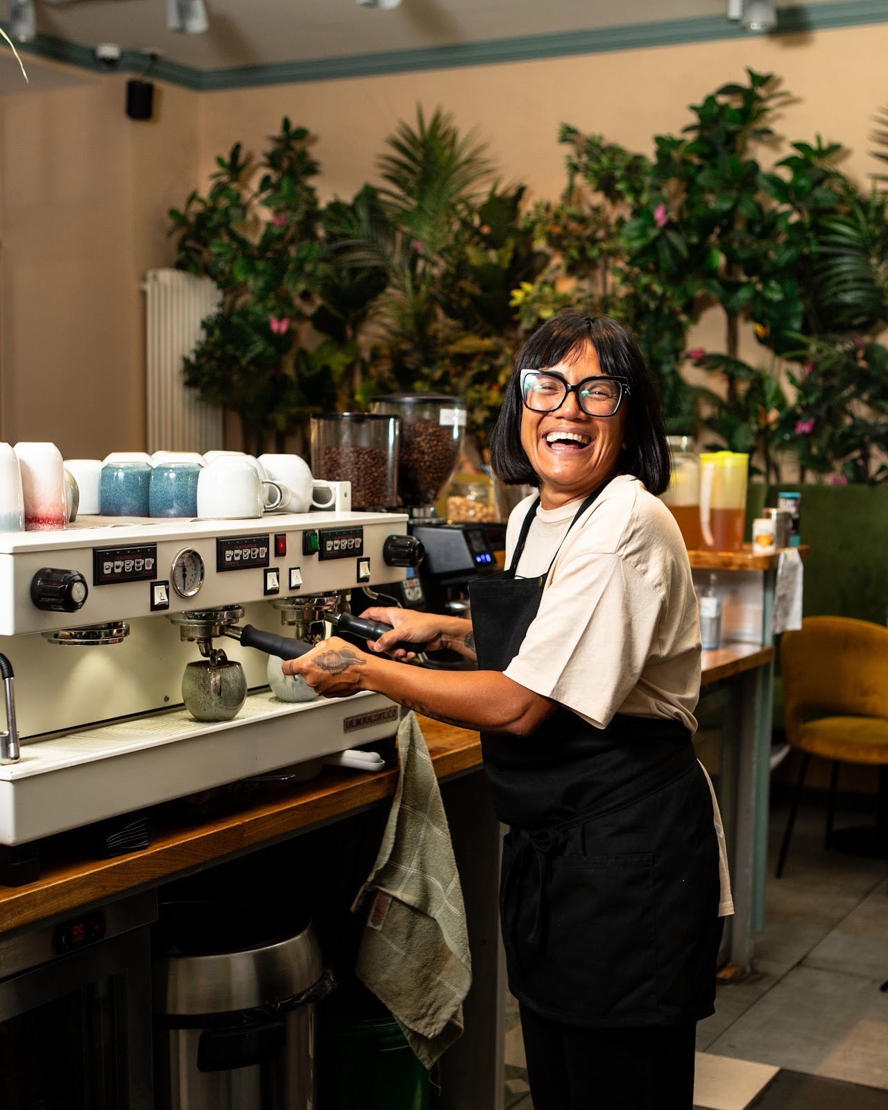 Innenansicht des Jederzeit Cafés mit einer langen, hellblauen Theke, an der ein Barista arbeitet, Regalen mit Pflanzen und Produkten sowie warmer Beleuchtung.