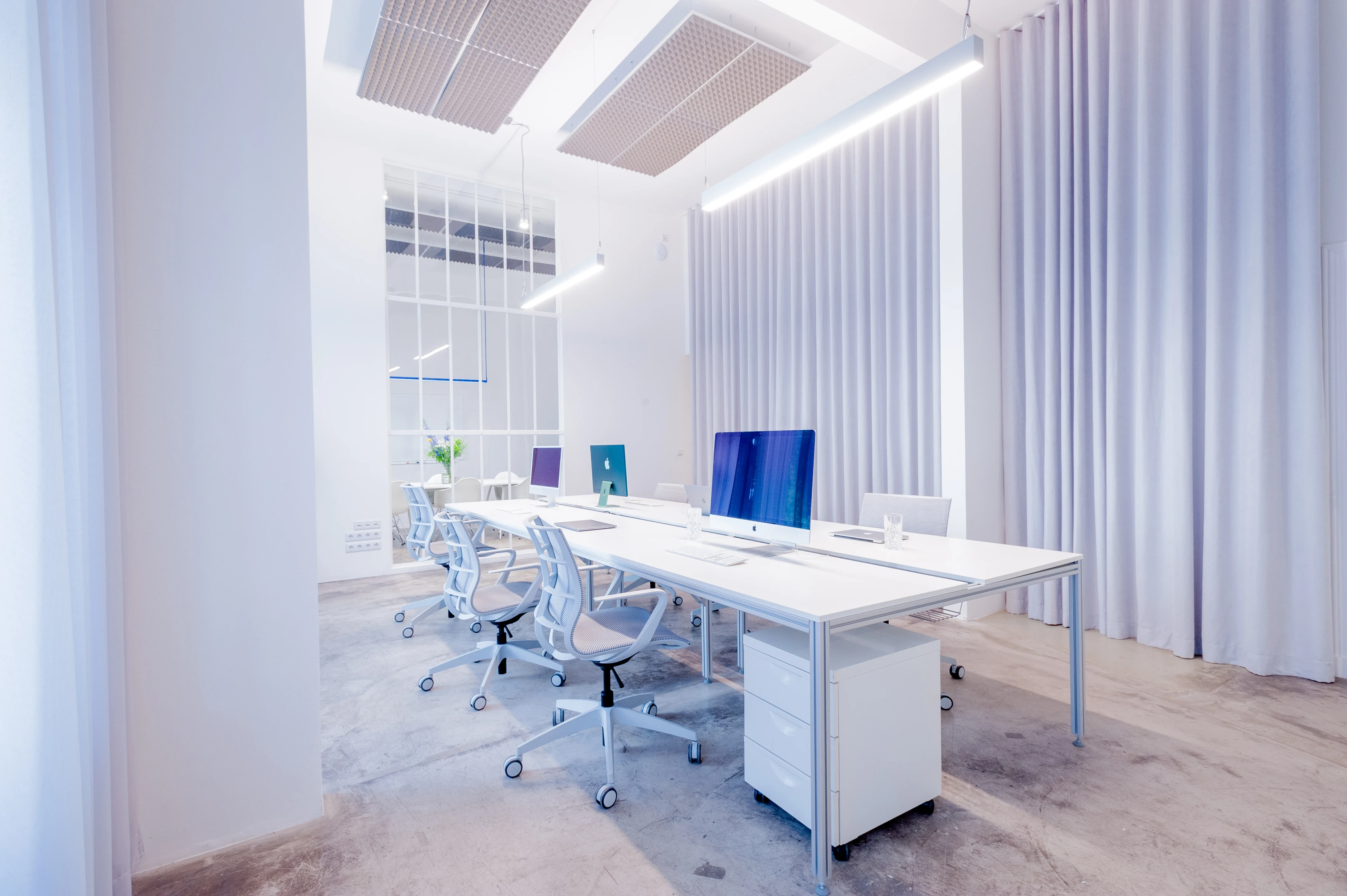 White workstations at BATTLE ROYAL with iMacs, white office chairs and rolling drawer units, framed by sheer wall-height curtains, linear pendant lights and perforated acoustic ceiling panels.