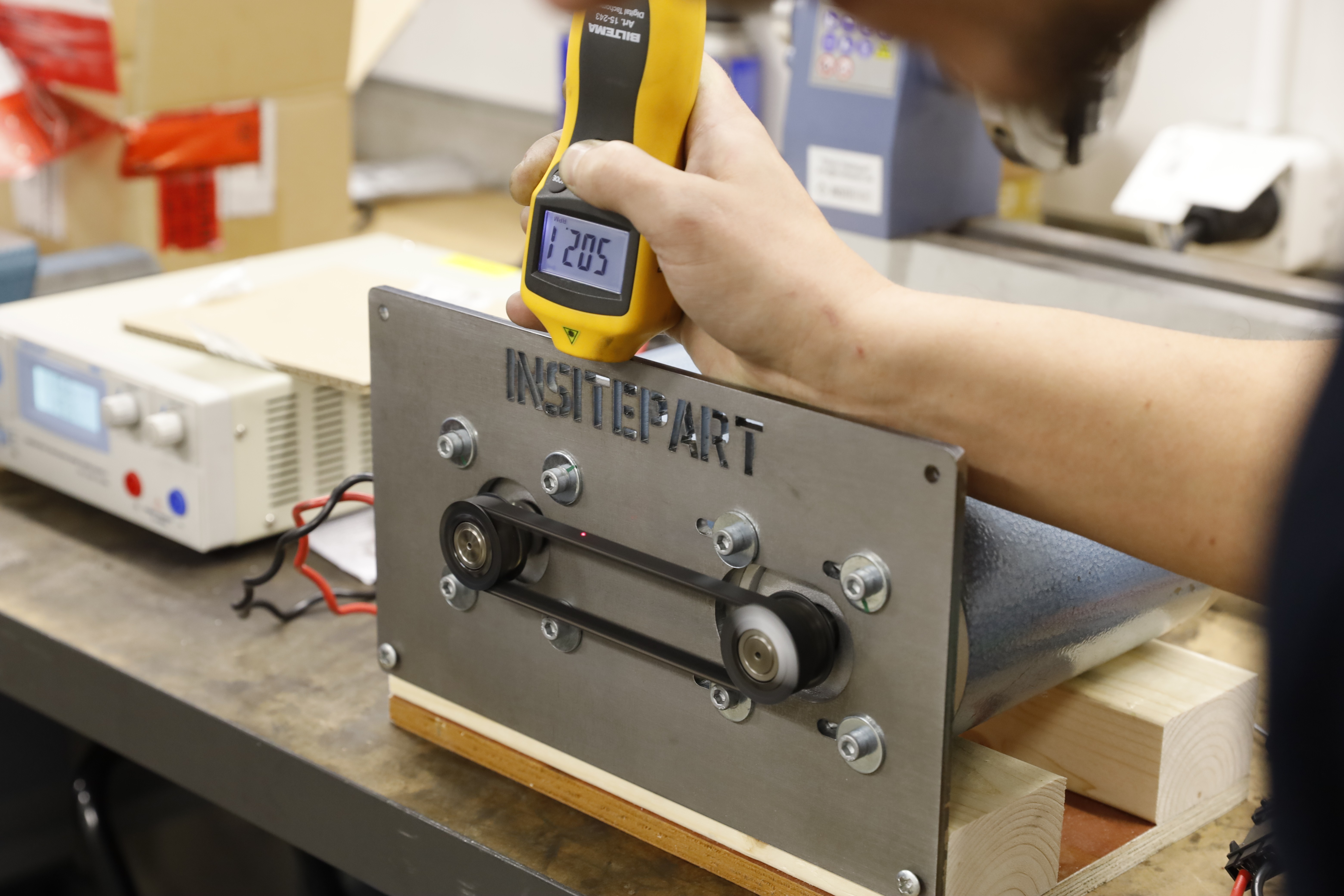 Person mäter temperaturen på en maskin med en termometer. Maskinen har remmar och en metallplatta.