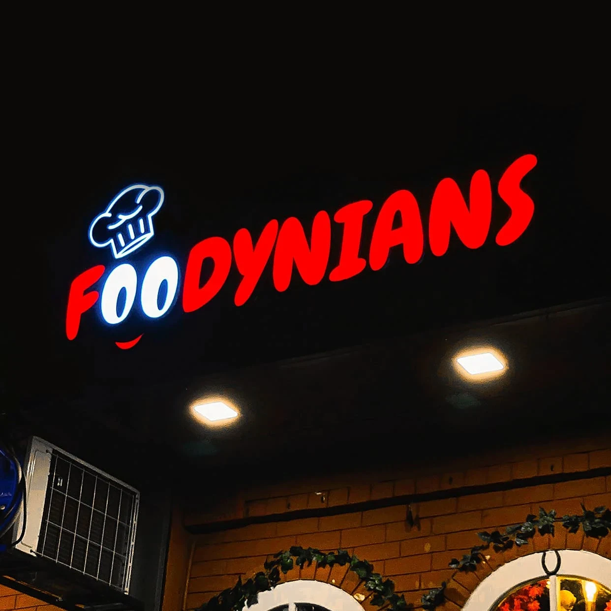 Neon “FOODNYANS” restaurant sign with a chef-hat logo glows above a brick facade, creating a lively, inviting nighttime atmosphere.