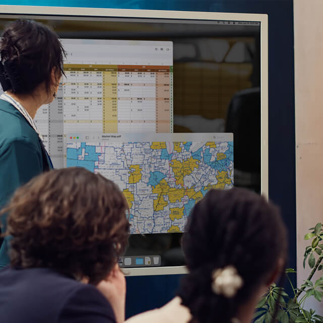 A person presents data on a large screen showing a spreadsheet and a color-coded map to an audience of three people.