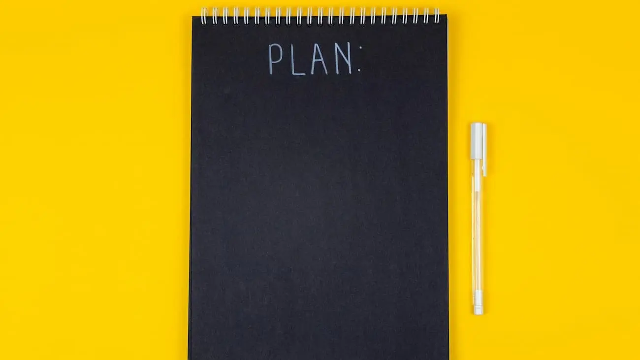 A simple yet striking image that captures the essence of meticulous planning, featuring a black spiral-bound notebook with the word "PLAN:" neatly written in white on its cover, accompanied by a transparent pen. This image, set against a vibrant yellow background, perfectly encapsulates the initial stage of organising a successful event, serving as a visual metaphor for the importance of a well-thought-out plan in the execution of a tournament. The clear, bold colours and minimalist design make this photo ideal for use in a blog article about event planning, offering a visual cue to readers about the significance of strategising and preparing detailed checklists in the realm of tournament organisation.