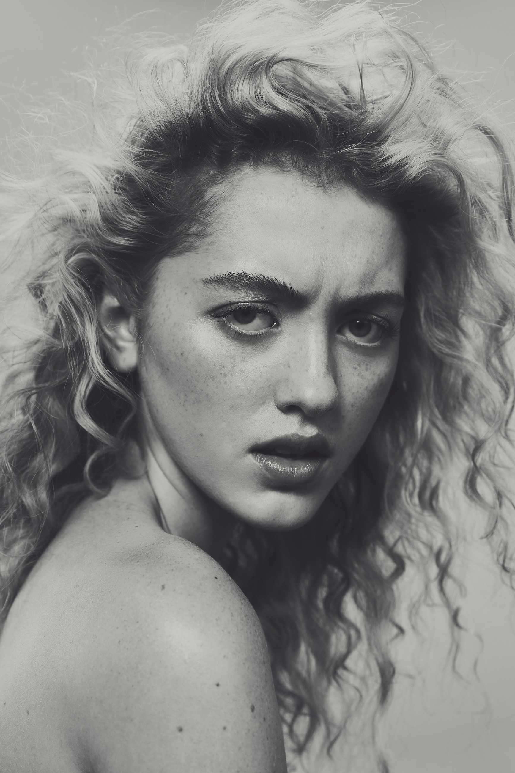 Black and white beauty portrait showcasing dramatic hair movement and natural freckles, with intense gaze and sculptural lighting creating contrast and depth.