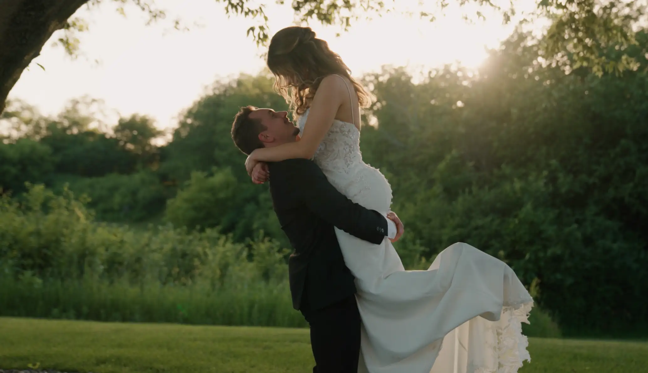 A Wisconsin couple in formal attire shares a joyful moment outdoors, the groom lifts the bride in a scenic sunset backdrop.