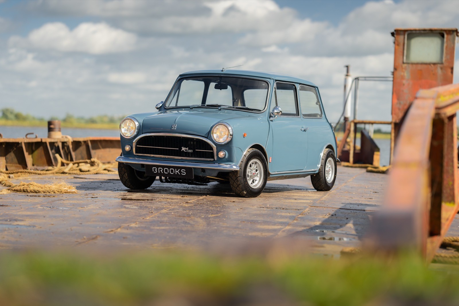 A vintage blue car parked near a wooden structure, with a cloudy sky in the background and grass in the foreground.