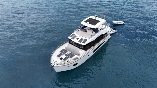 Elevated aerial bow view of the Cranchi T55 Eco Trawler looking aft, clearly showing the large foredeck solar panel array, flybridge solar panels, black panoramic superstructure glazing, side deck rails, and a RIB tender at anchor astern, highlighting the yacht's eco-trawler sustainable energy system on calm open water