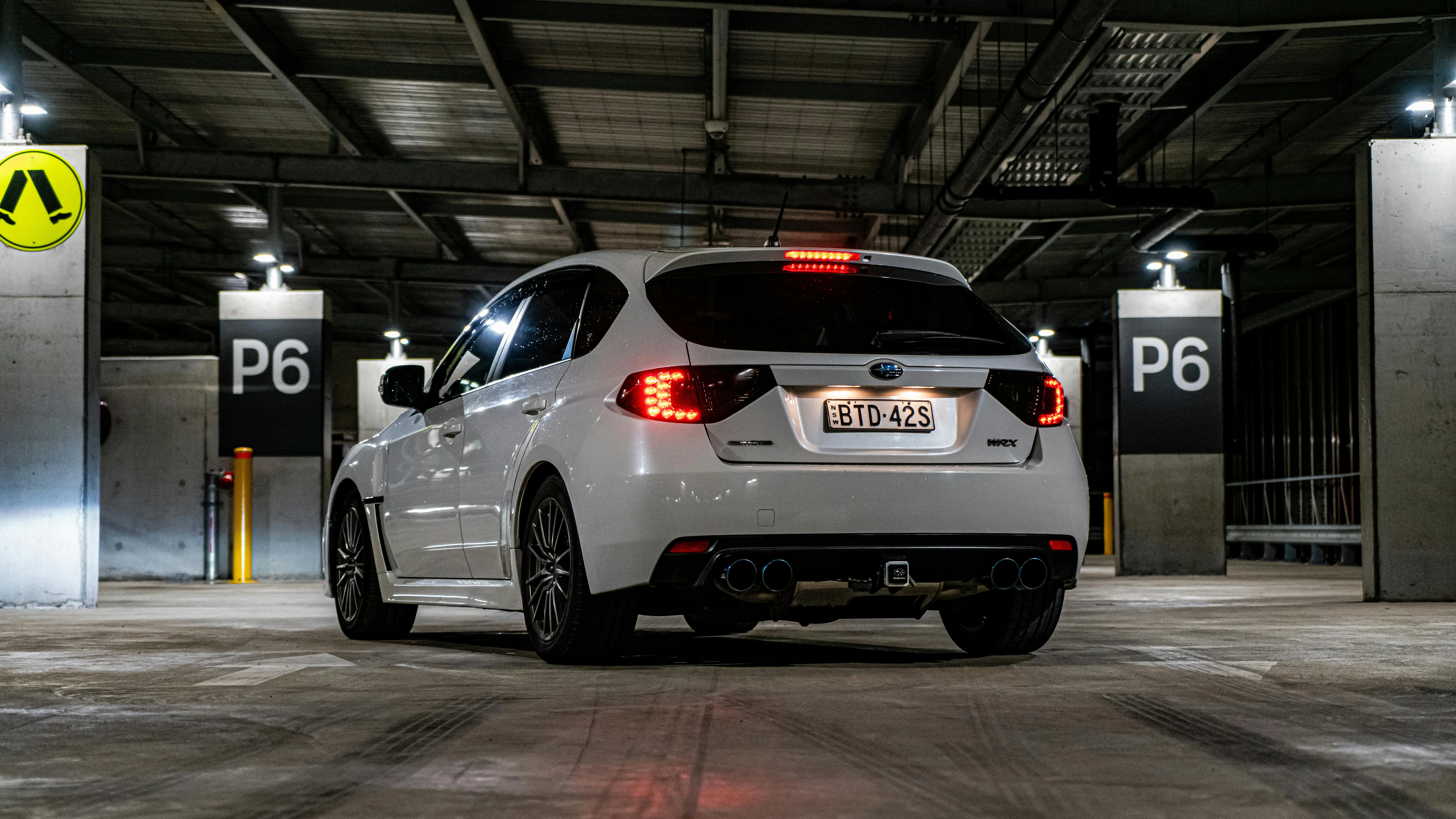 Subaru WRX blanche photographiée de l’arrière dans un parking souterrain. Les feux LED rouges, la quadruple sortie d’échappement et les jantes sportives soulignent le caractère agressif du modèle. L’environnement industriel, les poteaux numérotés et les éclairages artificiels créent une scène urbaine moderne et intense.