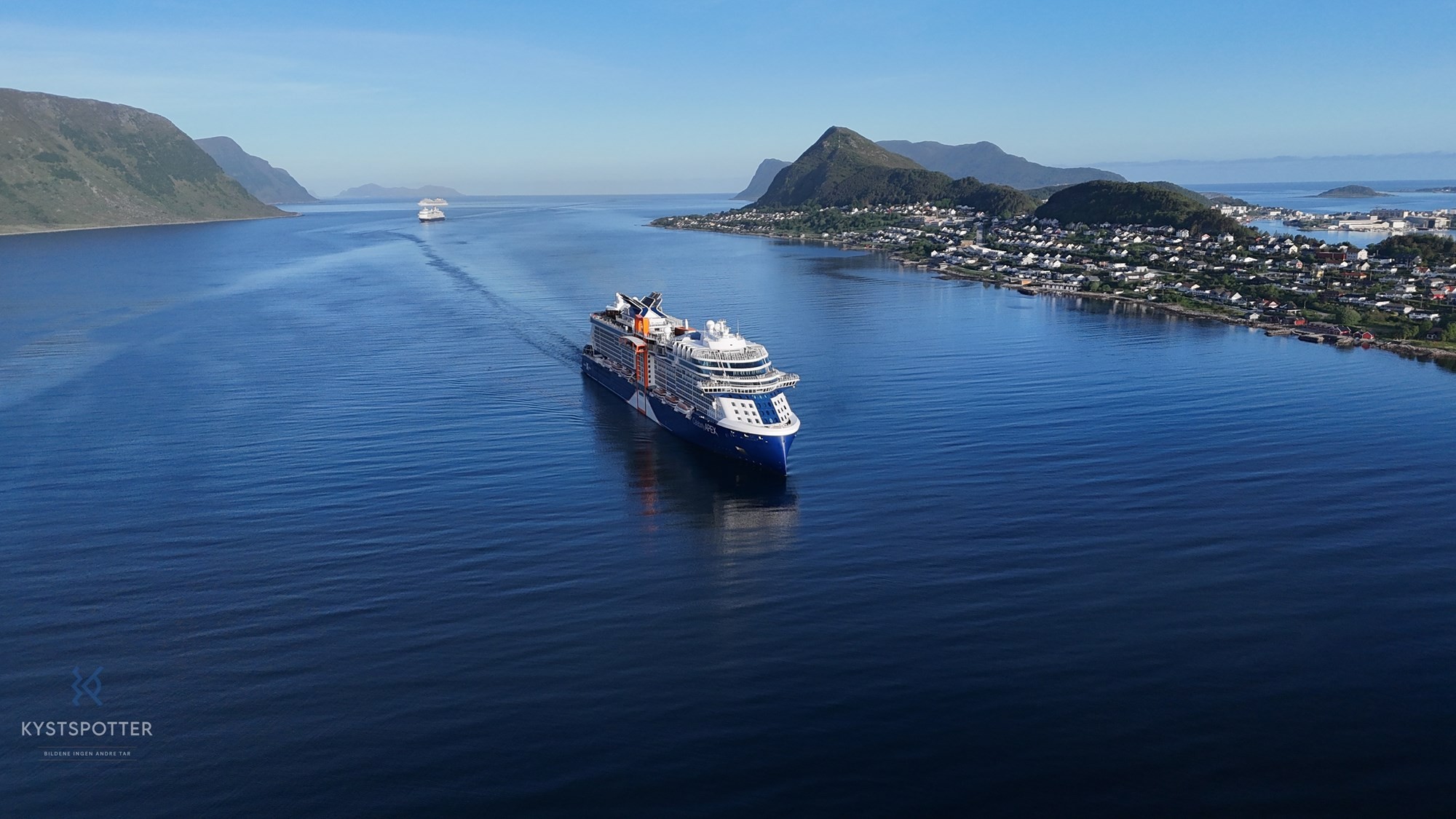 Aerial view of a cruise ship sailing on calm waters surrounded by mountains and clear blue sky.