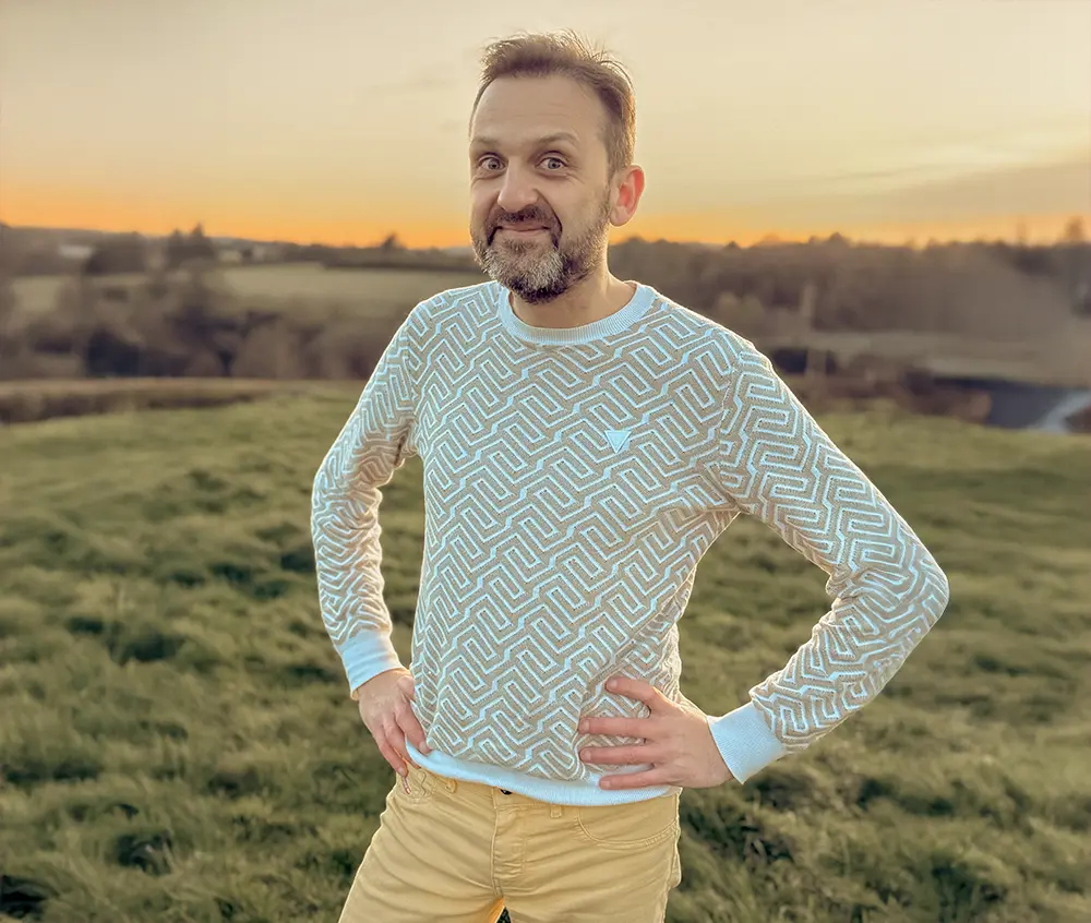 Confident man standing in a greenhouse surrounded by lush green plants, wearing a beige shirt and neutral-toned outfit.