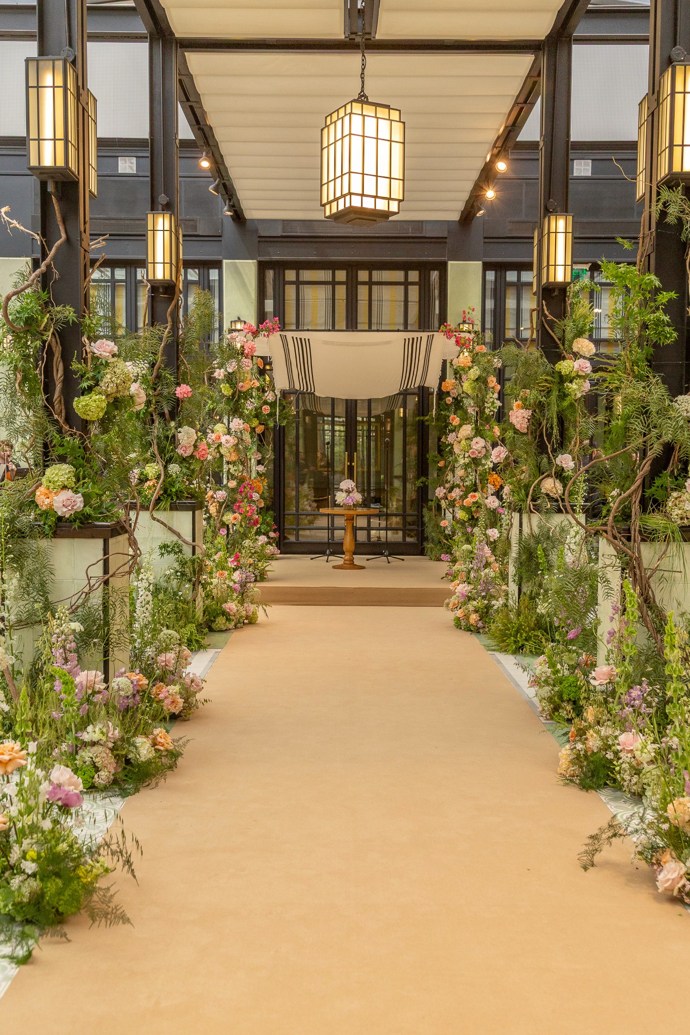 Camino al altar en salón de lujo con alfombra beige, estructuras botánicas altas y lámparas colgantes modernas.