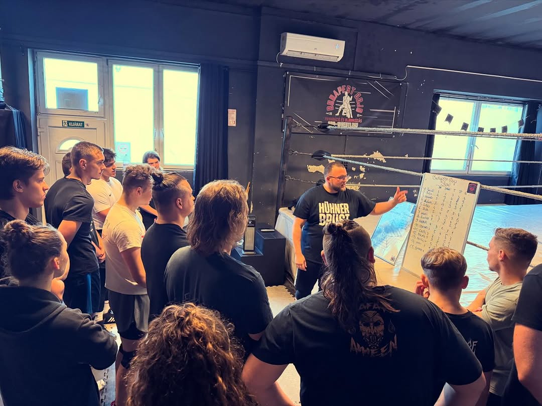 Three people in casual outdoor attire stack firewood into a rustic stone oven amidst an olive grove, with a sunlit house in the background, capturing an authentic rural lifestyle scene.