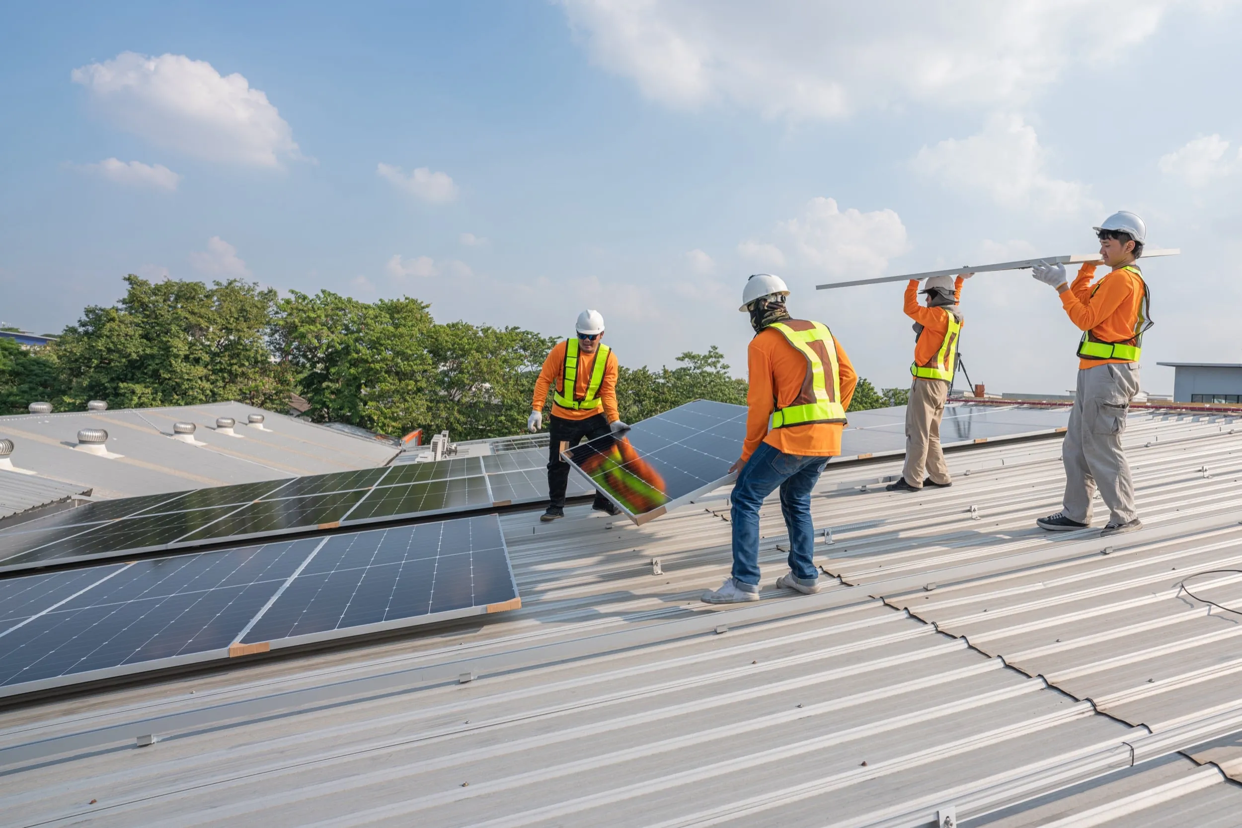 Vier Arbeiter in Sicherheitsausrüstung und orangefarbenen Hemden installieren ein großes Solarpanel auf dem Dach eines weißen Hauses vor strahlend blauem Himmel und machen damit auf nachhaltige Energie und Solartechnologie aufmerksam.