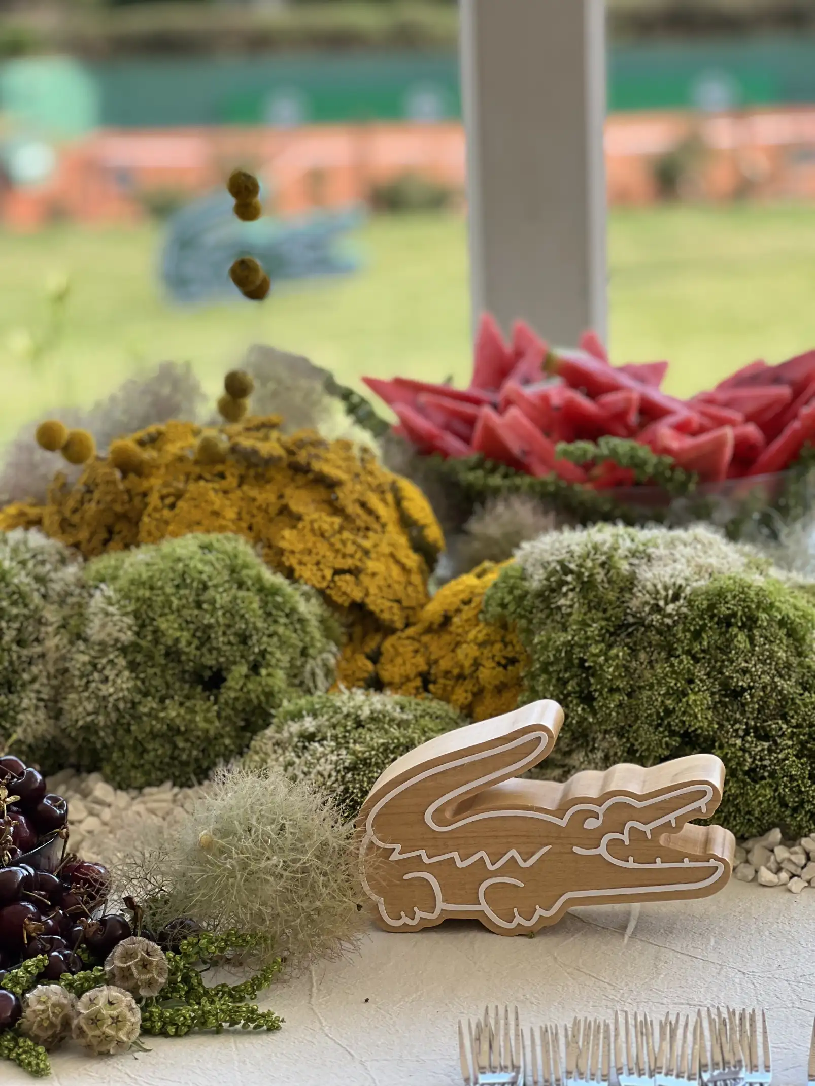 Centro de mesa con esferas florales verdes y amarillas acompañadas de frutas frescas, con un adorno de madera en forma del icónico logo de cocodrilo de Lacoste en primer plano.