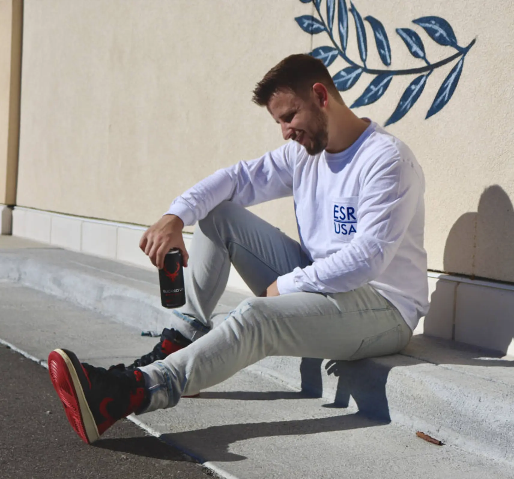 Man sitting on a curb in casual streetwear holding a Bucked Up energy drink can wearing a white ESR USA long-sleeve shirt and black and red sneakers.