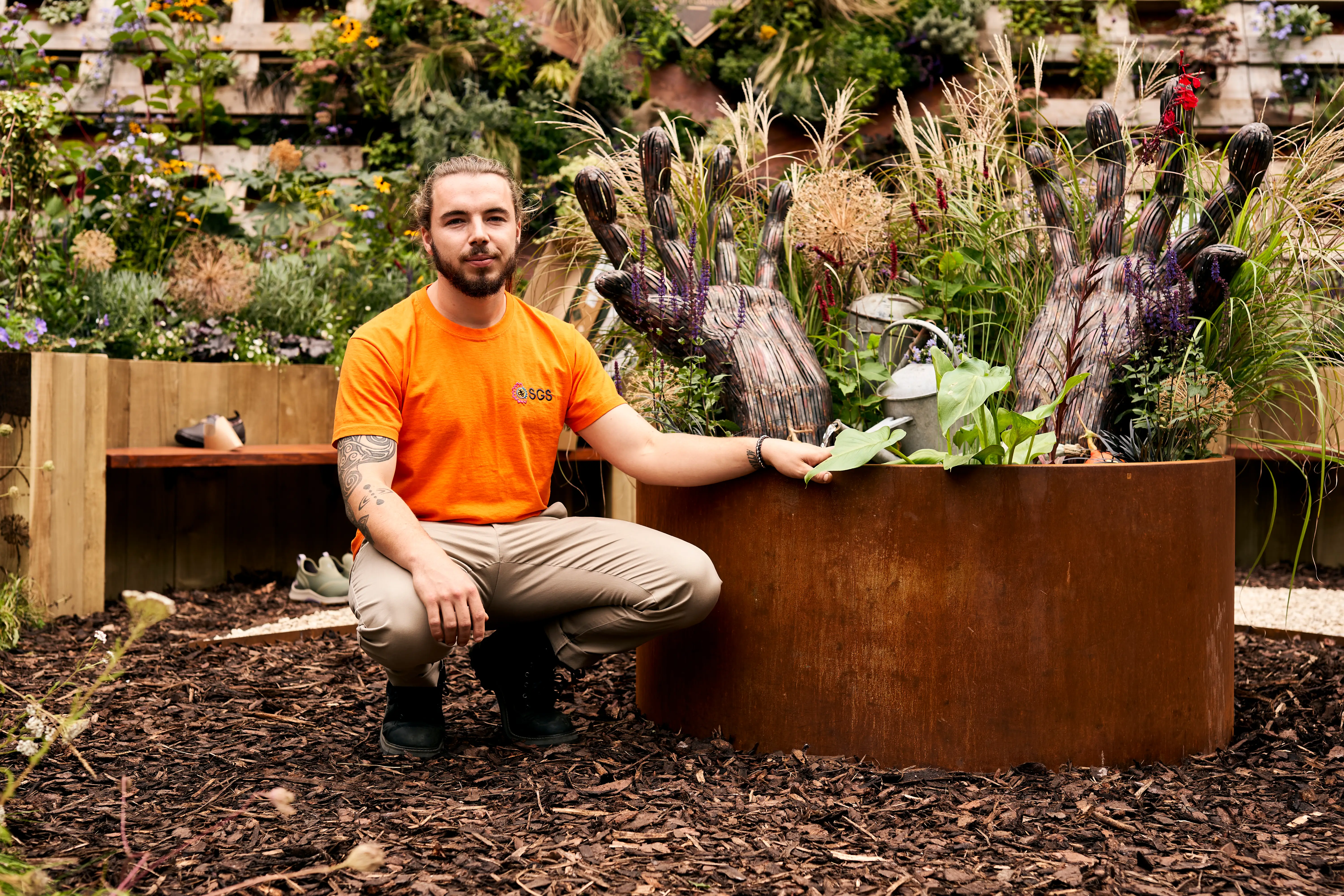 A person in an orange shirt kneels next to a planter, smiling in a garden filled with plants.
