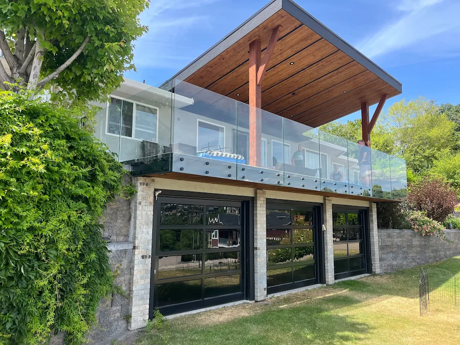 Frameless glass deck railing on an elevated balcony with a wood-beam roof, overlooking a residential property.