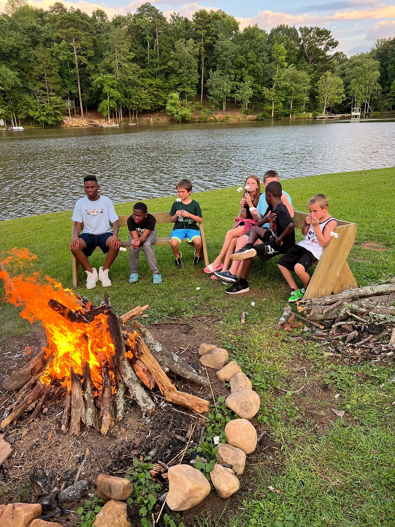 Group of campers sitting around a bonfire at dusk by a lake, enjoying s'mores.