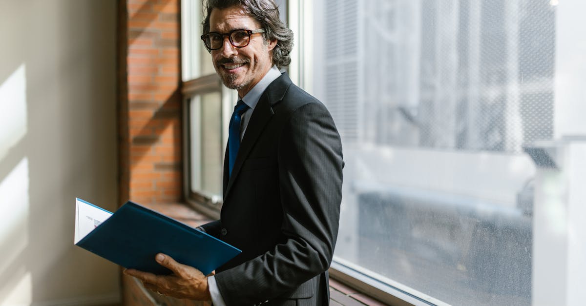 Smiling businessman in a black suit holding a folder by a window indoors.