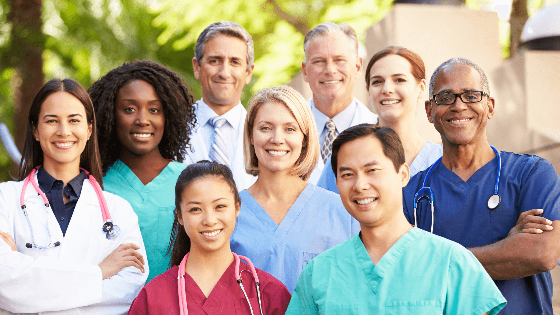Smiling group of diverse healthcare professionals in scrubs and lab coats standing together outdoors, representing a collaborative medical team.