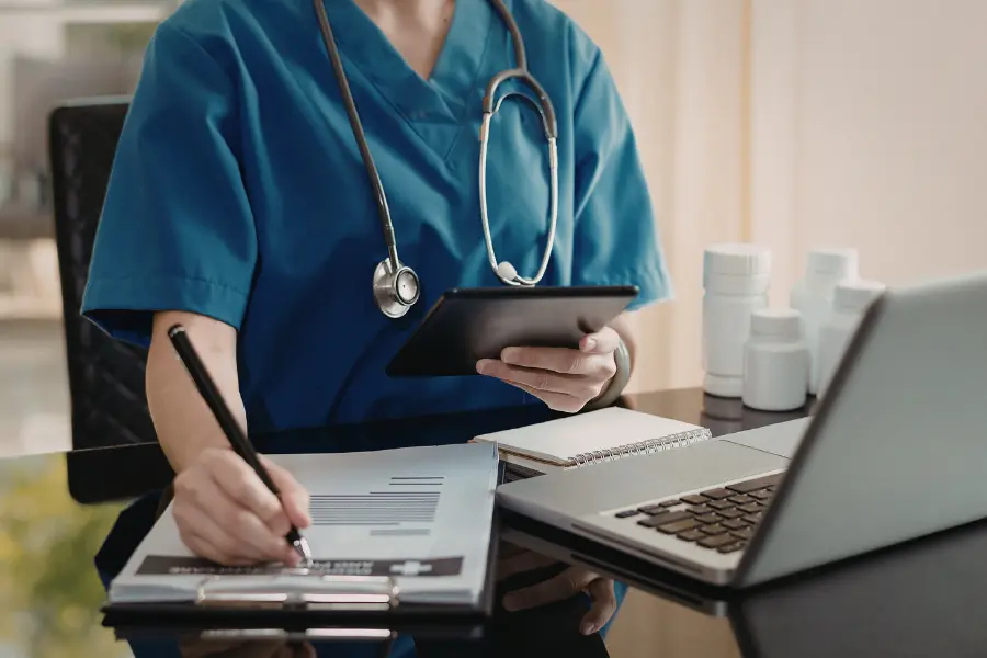 AI tools in healthcare assisting a nurse in blue scrubs who writes on a clipboard while referencing a tablet and laptop.