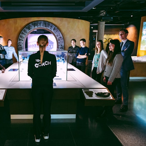 ਇੱਕ ਵਿਅਕਤੀ ਜਿਸਨੂੰ 'ਸ਼ਿਕਸ਼ਕ' ਦੇ ਤੌਰ 'ਤੇ ਚਿੰਨਿਤ ਕੀਤਾ ਗਿਆ ਹੈ, ਇੱਕ ਆਧੁਨਿਕ ਮਿਜੀਅਮ ਮੈਦਾਨ ਵਿੱਚ ਲੋਕਾਂ ਦੇ ਗਰੂਪ समਾਂਏ ਇੱਕ ਪ੍ਰਦਰਸ਼ਨੀ ਦੇ ਕੇਸ ਦੇ ਸਾਹਮਣੇ ਖੜ੍ਹਾ ਹੈ।