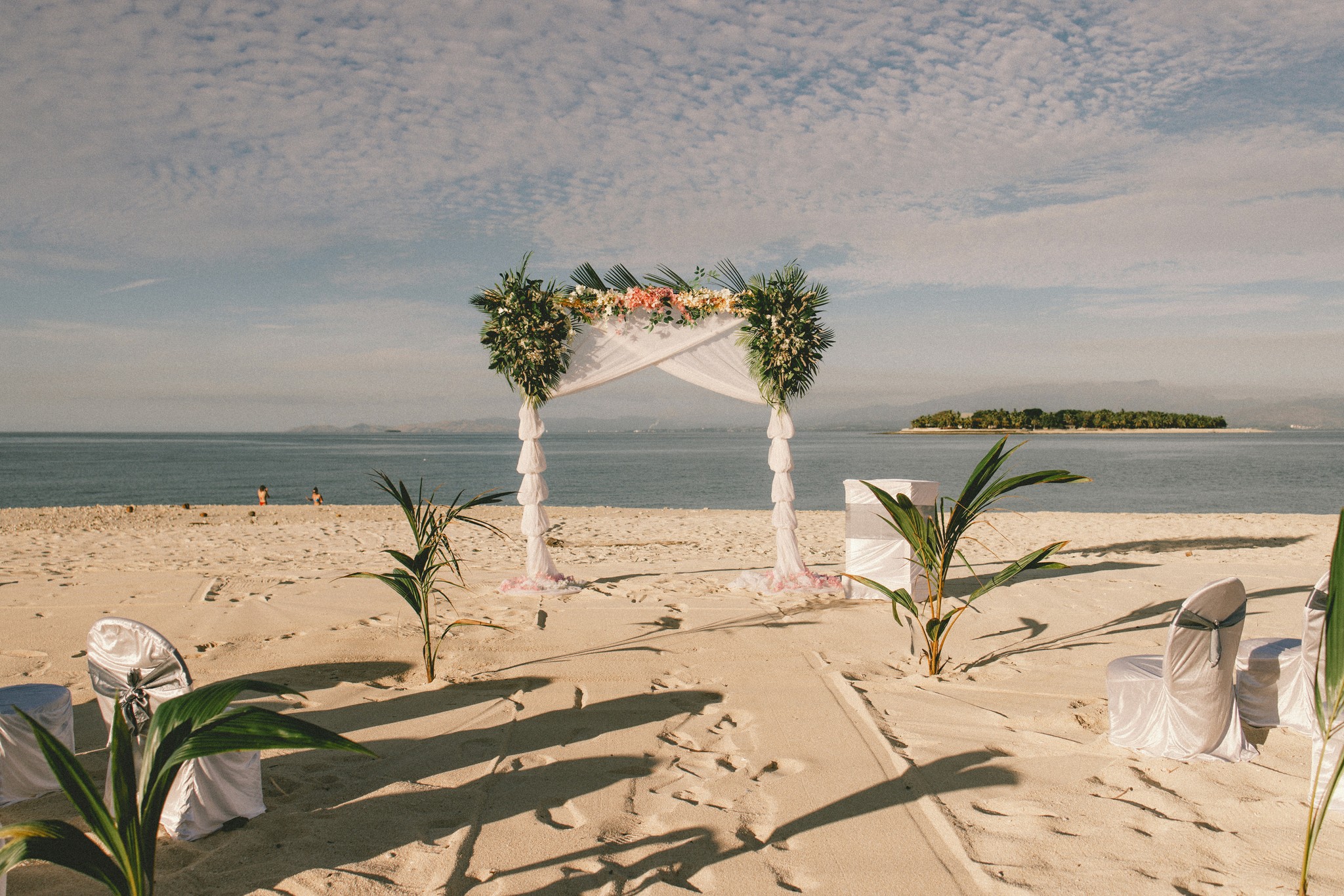 Tropical beach wedding arch decorated with white draping and palm fronds overlooking calm ocean waters and a distant island