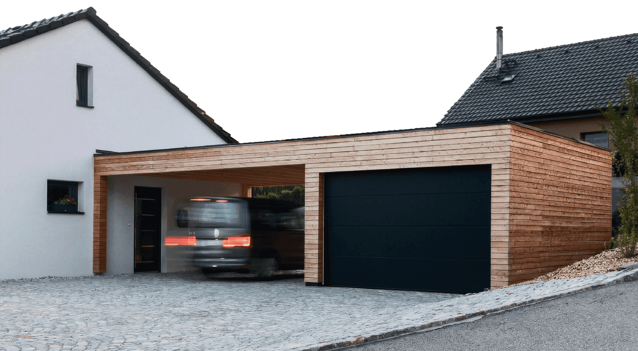 A modern residential exterior featuring a minimalist white house with a natural wood-clad garage and carport. A black sectional garage door is installed flush with the horizontal timber siding, set against a cobblestone driveway.