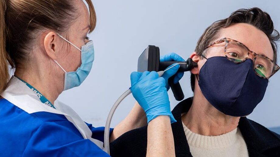 A healthcare worker examines a patient's ear using an otoscope. Both are wearing masks and gloves.