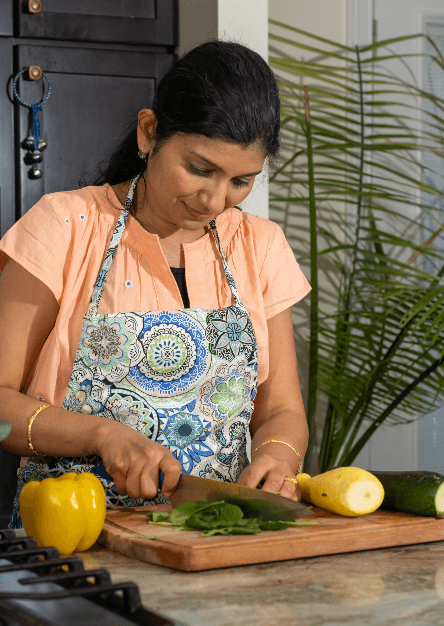 Sunita in a kitchen prepares Ayurvedic healing meals by chopping fresh green vegetables on a wooden board, surrounded by vibrant yellow bell pepper and zucchini, while wearing a floral apron.