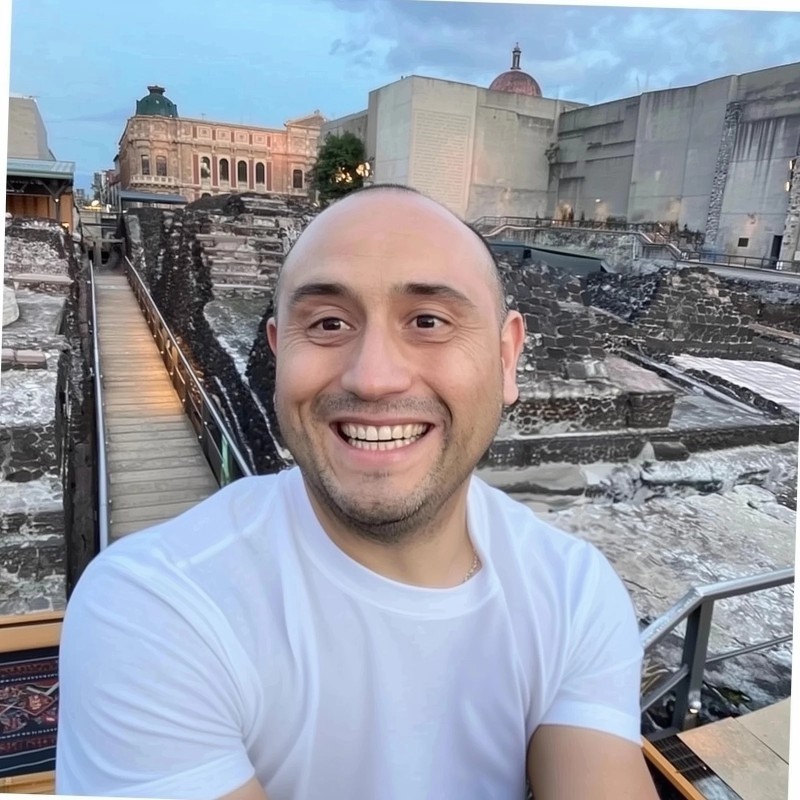 Young man with tousled brown hair and blue eyes, wearing a leaf-patterned shirt. Calm expression, indoor setting with blurred background.
