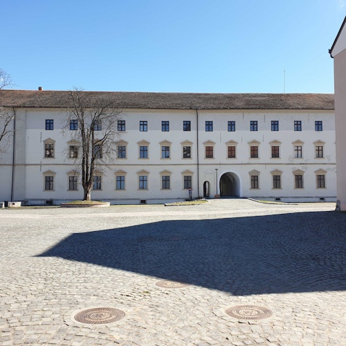 一座大型的白色建築,擁有多個窗戶和一個拱形入口,坐落在鵝卵石 courtyard 下,天藍藍的。