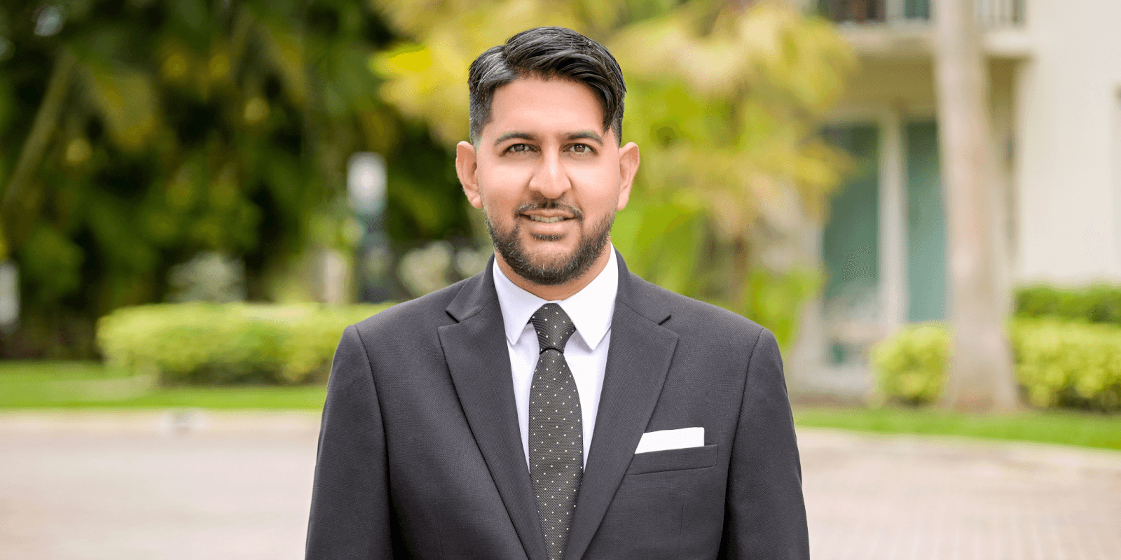 Andrew Roopchand, a luxury real estate agent, confidently poses outdoors in a dark suit, white shirt, and tie.