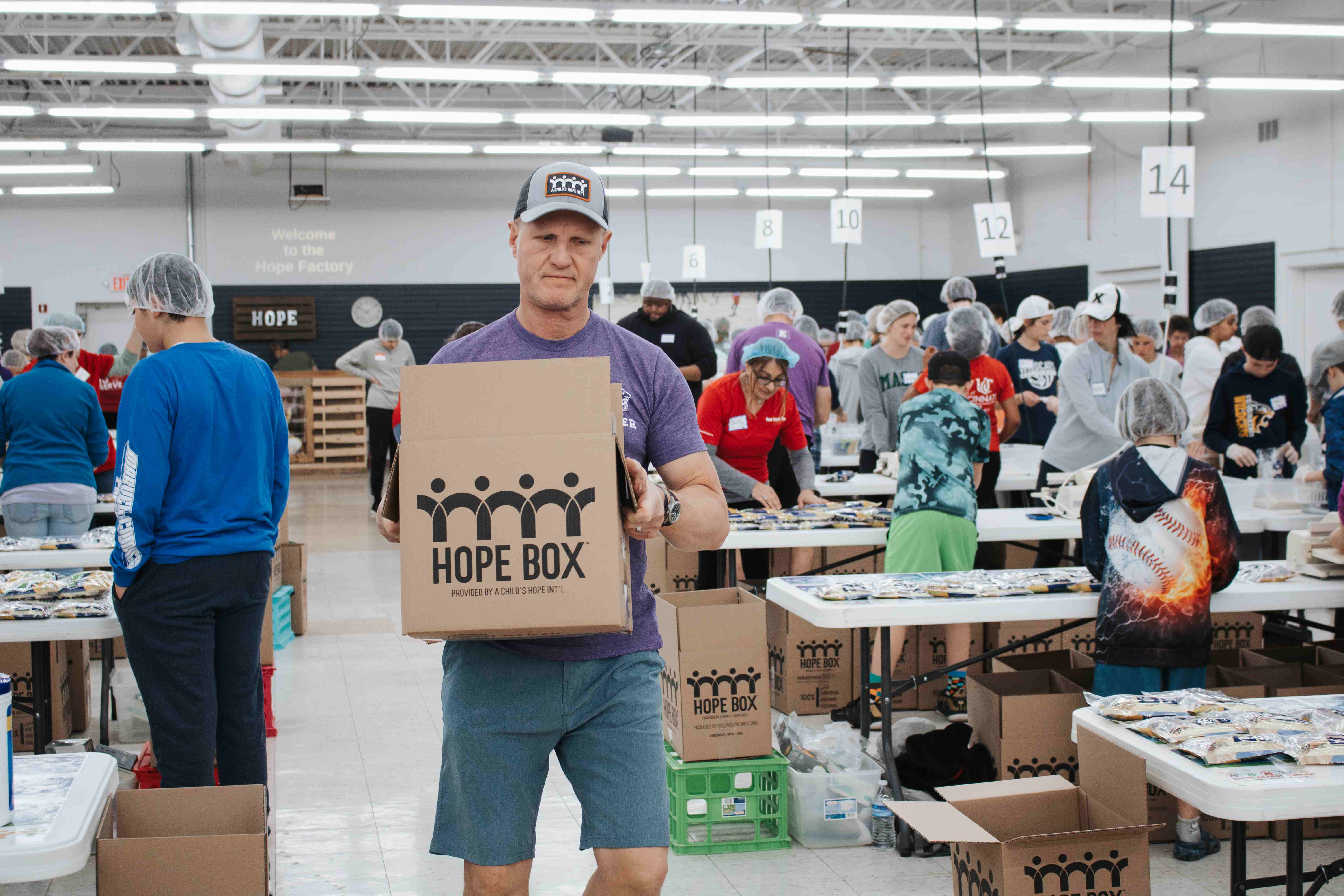 Man holding a Hope Box in a busy nonprofit meal-packing center, with volunteers working at tables in the background.